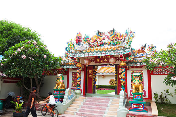 Nathon, Thailand - May 11, 2014: Entrance to chinese styled temple Hainan in Nathon on Ko Samui. From left side a Thai woman is walking to temple. In front of her is a girl on bicycle. On top of gate are dragons. Behind is huge mandarin warrior statue.