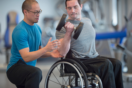 A disabled man sitting in a wheelchair is working out his arms at the gym by lifting dumbbells. His trainer is helping him with his form.