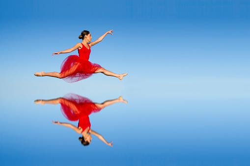 Side view of young woman in red dress dancing ballet against of blue background. Reflection. Gradient