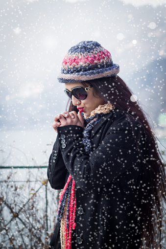 Young woman is shivering while snowing, in the background, a lake and a hill landscape. She is wearing dark sunglasses, black coat, colorful hat and scarf. shallow depth of field, writing space