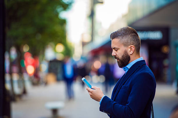 Hipster manager holding smartphone, texting outside in the stree