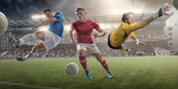 Composite image of male soccer players in action – football player in blue shirt in mid air kicking ball, football player in red shirt dribbling ball, goalkeeper in mid air making save. Action takes place on a generic outdoor floodlit soccer stadium full of spectators under dramatic stormy sky at sunset. 