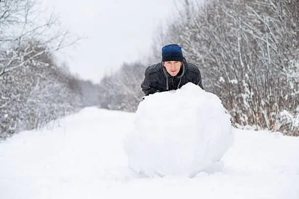 Boy rolls a big snowball to build a snowman in winter day