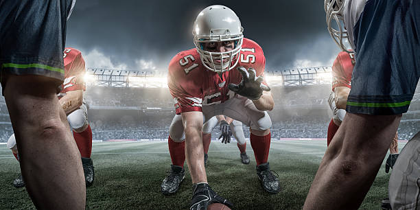 American Football Center in Offensive Line About to Snap Ball Close up image of an American Football player in center position in an offensive line formation with teammates. The athlete in a number 51 shirt is wearing eye black and looking at the camera, about to snap the ball. The players are playing in a generic outdoor floodlit stadium full of spectators. offensive line stock pictures, royalty-free photos & images