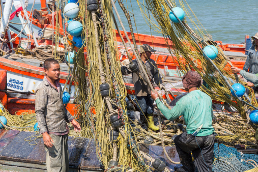 Ang Sila, Thailand- January 15, 2014: Fishermen cleaning the nets on colorful Thai trawler. Three fishermen are arranging the yellow fishing nets with blue floats. Sea in the background.