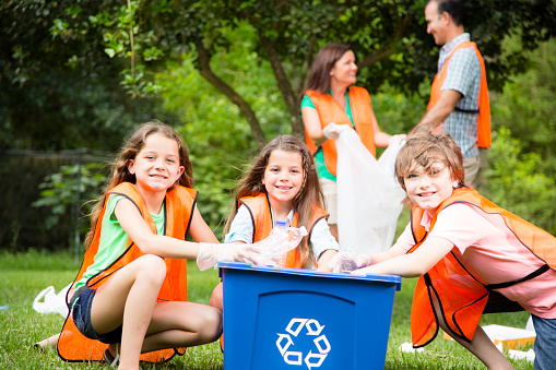 Community volunteers clean up their local park. Family recycles trash.  Children are in the foreground with a recycling bin as the other family members pick up garbage in the background with garbage bags. Father, mother and three children all wear safety vests.  Environmental conservation.  Nature.