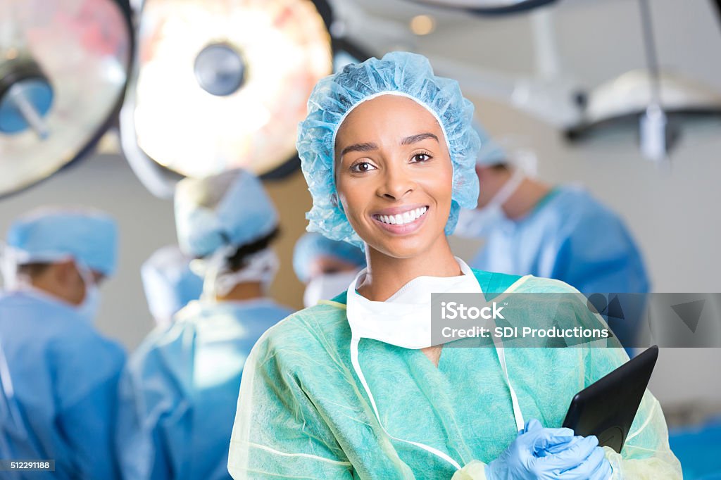 Young surgeon prepares for surgery Pretty young African American female surgeon prepares for surgery. She is wearing surgical gown, surgical mask and hair net. She is holding a digital tablet. a team of surgeons are operating in the background. Surgery Stock Photo