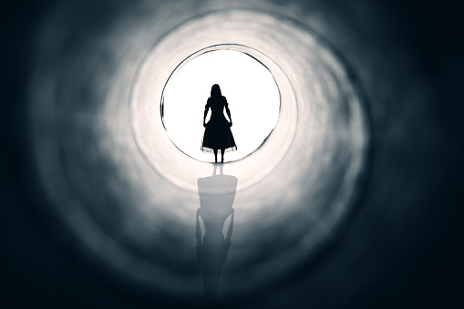 A conceptual image of a woman in a dress facing the light at the end of a tunnel.