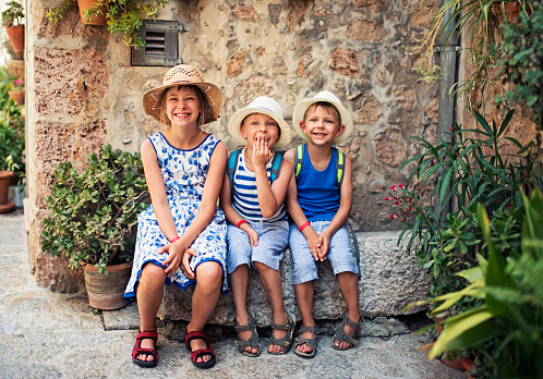 Three kids tourist resting in the beautiful mediterranean town. Little boys are wearing backpacks and hats. Theirs sister is wearing sundress. Kids are sitting on stone bench and laughing.