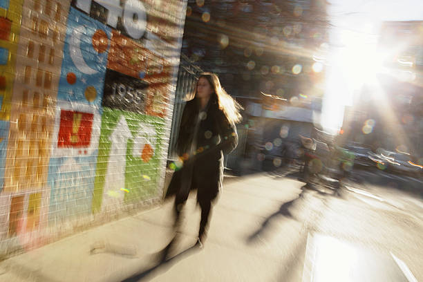 Defocused image of sun rising down NYC backstreet New York, NY, USA - February 26, 2016: Defocused blurred image of the sun rising down Rivington Street on the Lower East Side of New York City as an unknown woman walks down the sidewalk. A colorful art mural is painted on the wall of P.S. 140 Nathan Straus public school which was built in 1958. In the background are tenement buildings and high rise low cost apartment buildings. The rising sun is causing lens flare and the long exposure is causing the image blur. rivington street stock pictures, royalty-free photos & images
