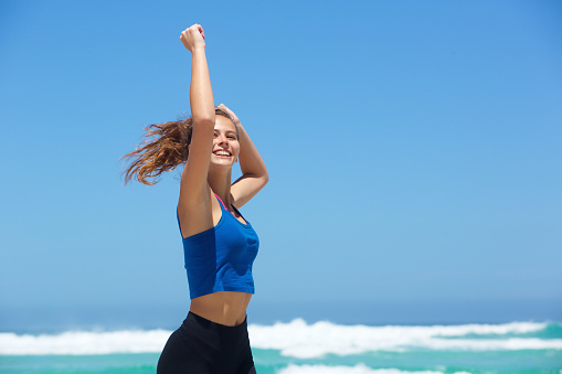 Side portrait of a fit young woman running by the sea with arms raised