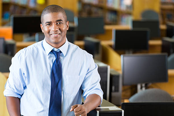 African American mid adult male high school teacher is standing in computer lab in school's library before class. He is looking at the camera and smiling confidently. His is wearing a button down shirt with the sleeves rolled up and a blue tie. Desktop computers and bookshevles are in the background.