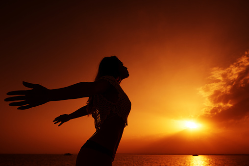 side view of silhouette of a woman with arms raised at sunset, feeling free, sea side in background, tranquil moment.