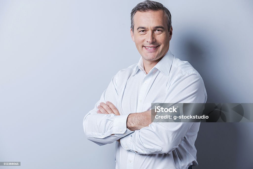 Confident mature man. Portrait of confident mature man in shirt looking at camera and smiling while keeping arms crossed and standing against grey background Men Stock Photo