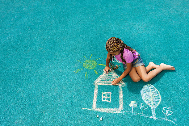 Nice looking black girl drawing house sun and trees with chalk on playground