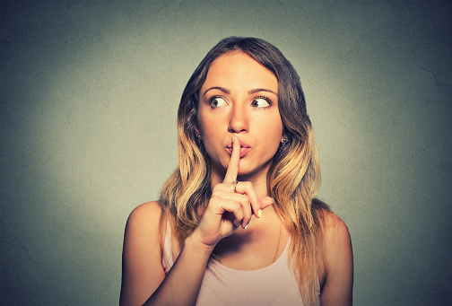 Closeup portrait secretive young woman placing finger on lips asking shh, quiet, silence looking sideway isolated gray background. Human face expressions, sign emotion feeling body language reaction