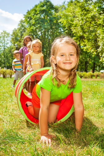 Group of children play with tube on the lawn with nice Caucasian girl crawling out of pipe with smile and her friends standing in the line