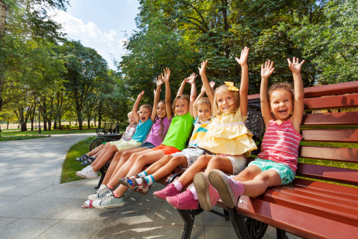 Large group of children sitting on the bench in the park with lifted hands and happy cheerful expression lifting hands