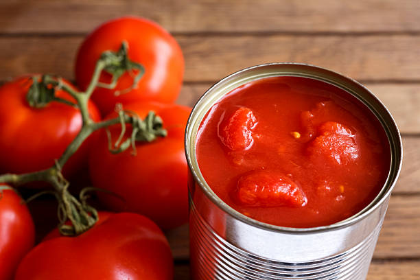 Open tin of chopped tomatoes. Open tin of chopped tomatoes with whole fresh unfocused tomatoes behind. Wood surface. preserved stock pictures, royalty-free photos & images