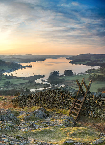 View of a sunrise over Windermere in the Lake District A picturesque view from Loughrigg Terrace looking across Windermere lake in the Lake District as the sun rises and casts early light on a stone wall and stile english lake district stock pictures, royalty-free photos & images