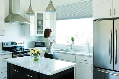 A woman stands looking out a window in a modern kitchen. She is drinking coffee.