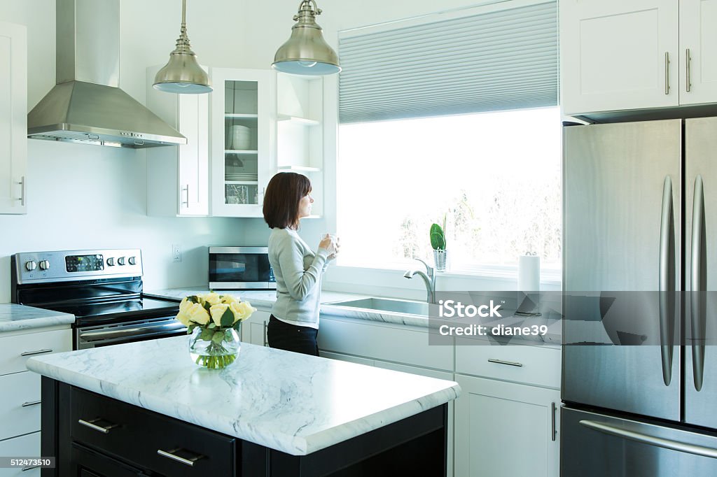 Woman Drinking Coffee in a Modern Kitchen A woman stands looking out a window in a modern kitchen. She is drinking coffee. Kitchen Stock Photo