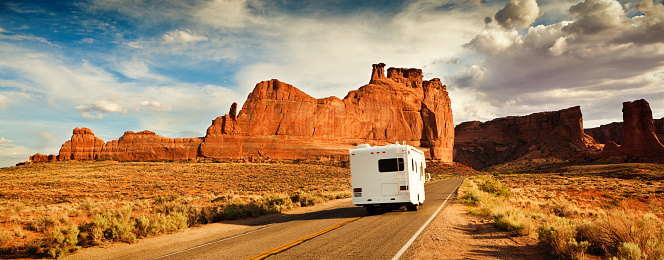 Motor home Camper on a vacation road trip, tourists exploring the American Southwest, the Arches National Park in Utah, USA. The well equipped motor home, traveling a famous scenic highway. Dramatic sky clouds and unique rock formations define the landscape of a popular western recreational travel destination.