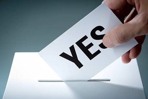 Voter's hand holding a YES vote ballot, inserting the vote card into the ballot box. Concept image of democracy and the election process for corporate election, board election, and governmental or business policy decisions. Voters making their selection and voting through a proxy ballot. Photographed in horizontal format.