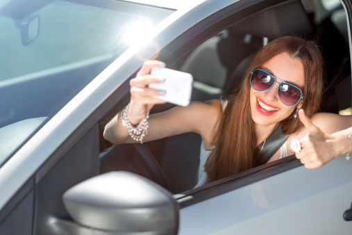 Smiling young woman taking selfie picture with smart phone camera outdoors in car