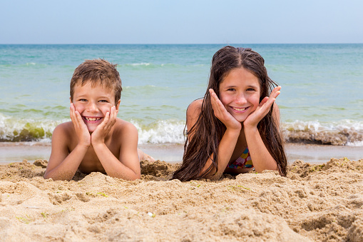 Two smiling kids on the beach lying down together near coastline