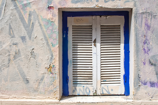 Blue framed window with white closed shutter. Wall of the house with partly covered graffiti. Plaka, Athens, Greece.