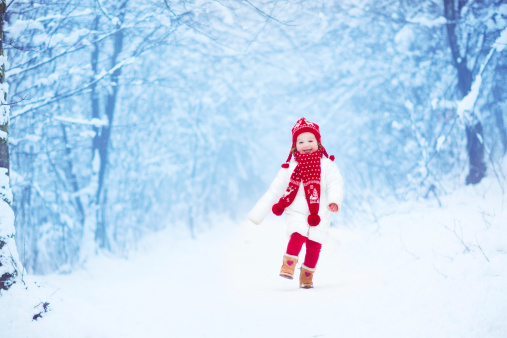 Happy laughing toddler girl wearing a white down jacket and red knitted hat and scarf playing and running in a beautiful snowy winter park on Christmas day