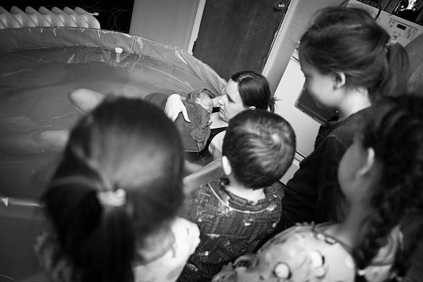 Siblings Meeting Newborn Brother After Home Water Birth Black and white image of a young mother embracing her newborn son after giving birth at home, while her other children see their little brother for the first time. water birth stock pictures, royalty-free photos & images