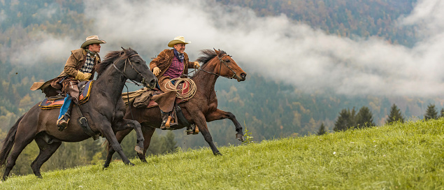 Two horseback riders in cowboy clothes riding across a field in a foggy forest.