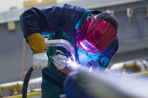 Industrial worker with protective mask welding inox elements in steel structures manufacture workshop.