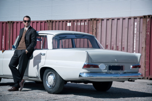 Retro 60s fashion business man wearing grey suit and black sunglasses standing against classic car.