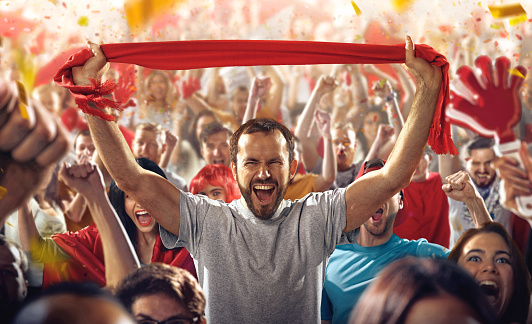 :biggrin:On the foreground a group of cheering fans watch a sport championship on stadium. In the centre a man shouting while holding a red scarf above his head. Everybody are happy. People are dressed in casual cloth. Colourful confetti flies int the air.