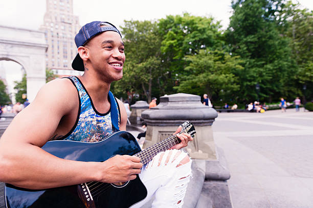 This is a color, royalty free, stock photograph of a smiling young Black man in his 20s playing the acoustic guitar outdoors on a summer day in NYC's Washington Square Park. Photographed  with a Nikon D800 DSLR camera.