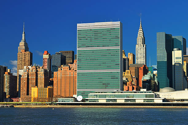 New York, NY - July 27, 2021: The United Nations General Assembly Building at UN Headquarters in East Midtown Manhattan, NYC. The building is seen from the East River. The UN Flag flies next to the building and sculptures from the UN Collection can be seen.