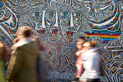 Berlin, Germany - April 17, 2014: Tourists walk along the East Side Gallery, an international memorial for freedom with paintings by artists from all over the world on one of the longest remaining sections of the Berlin Wall. This section was painted by Schamil Gimajew, and is entitled 'Worlds People'. 