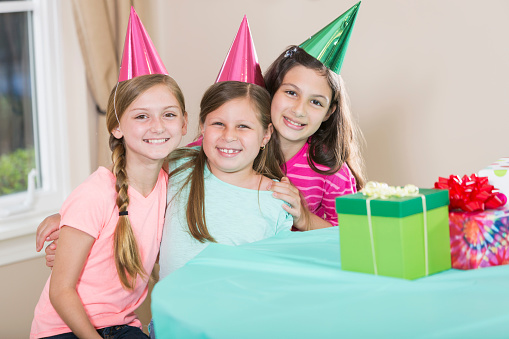 Three multi-racial girls having fun at a birthday party. They are posing for the camera, smiling together, wearing party hats. There are gifts on the table in front of them.