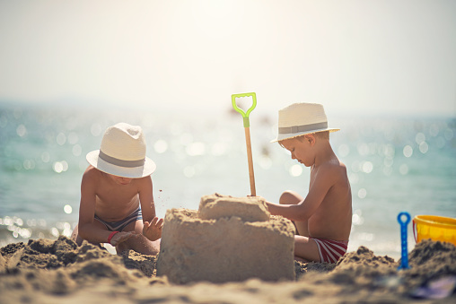 Brothers wearing funny fedora hats having fun building a sandcastle on the beautiful majorcan beach beach. The boys are aged 4. Sunny summer day.