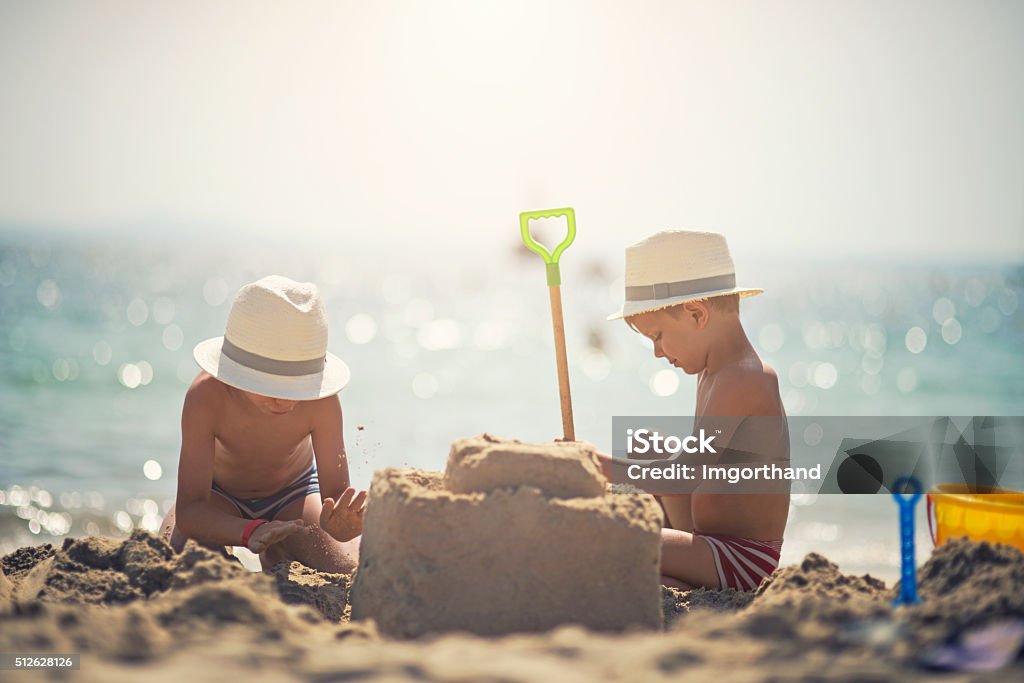 Two brothers building a sandcastle on beautiful beach Brothers wearing funny fedora hats having fun building a sandcastle on the beautiful majorcan beach beach. The boys are aged 4. Sunny summer day. Child Stock Photo