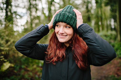 A young adult female with red hair puts on a hat during a hike. She is smiling with her hands on her head as she standing in the forest.