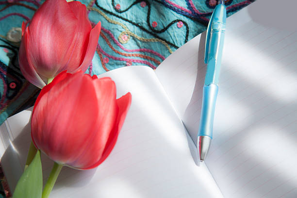 An open, blank notebook or diary with a cross bookmark lies on a table with pen.  A woman is about to begin her daily devotional time as she prepares to record her thoughts on the journal's blank pages.  No people.