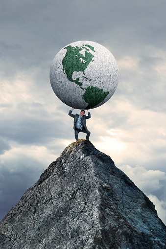A business stands on a mountain top as he struggles to lift a large globe rotated towards the Western Hemisphere above him.  Stormy skies hover in the background.