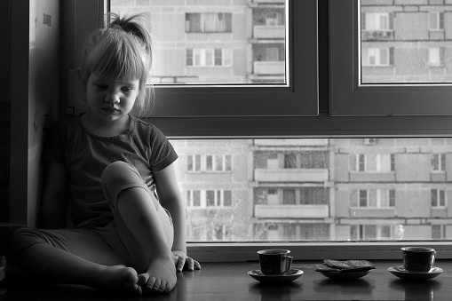 Sad child sits on a window sill of the window. Near two cups of tea. The concept of children's loneliness, the dream of a family, the loss of parents. Dirty window. Black and white