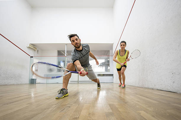 Below view of young man and woman playing squash. Low angle view of young couple exercising racketball on a court. Focus is on man reaching for the ball. squash sport stock pictures, royalty-free photos & images