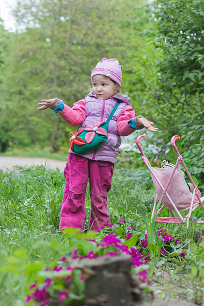 Little girl with pink toy stroller is making helpless shrug gesture near purple primula flowerbed