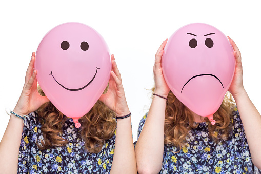 Two women holding pink balloons with happy and angry facial expressions for their heads isolated on white background. Two teenage girls hold inflated pink balloons with drawn facial expressions. Girls holding pink balloons in front of head with smiling, happy and angry, irritated facial expression. The balloons replace the faces of the women. The eyes and mouths are outstanding black. The girls are unrecognizable, they are anonymous. With isolated white background.